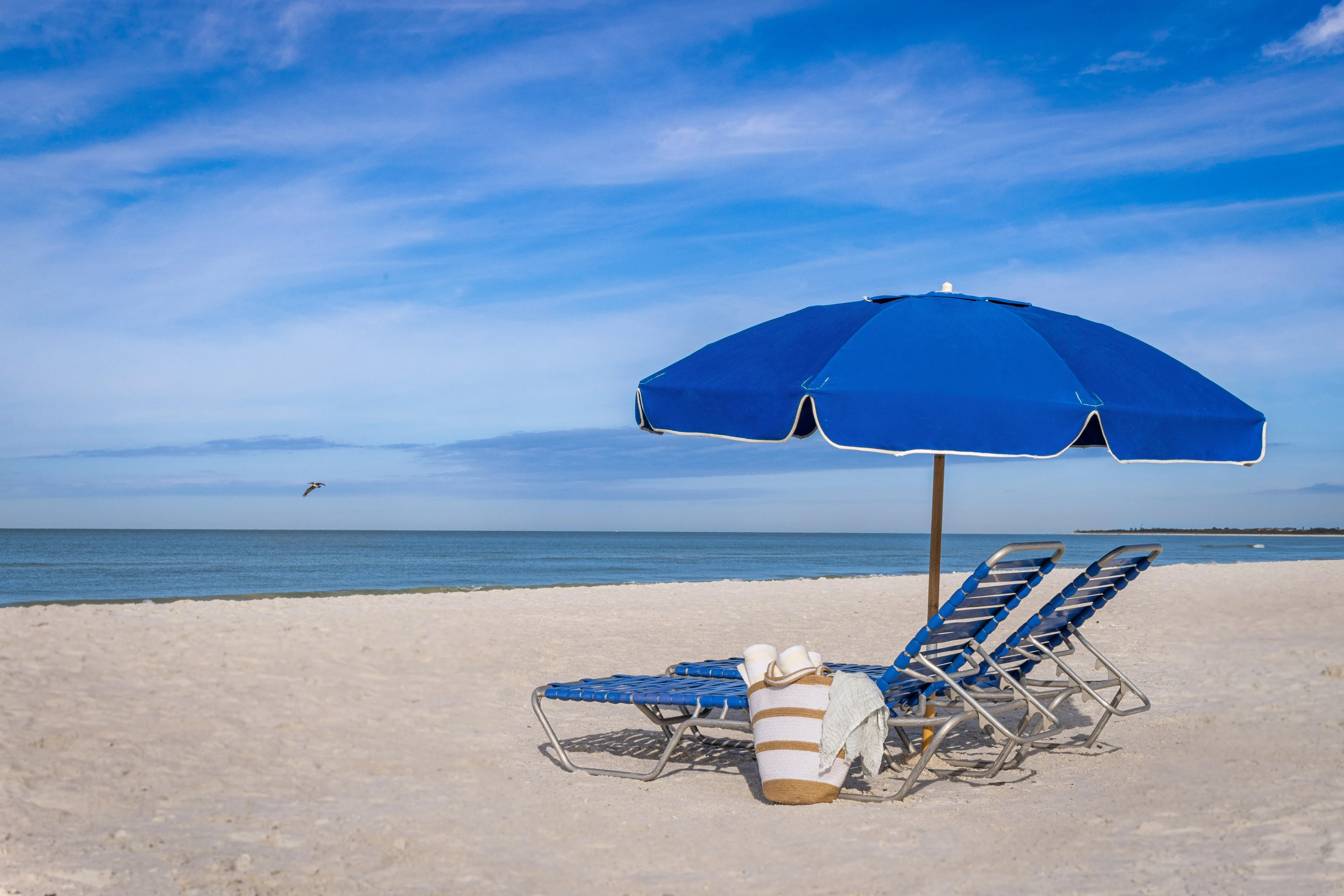 A row of blue lounge chairs sits beneath a large blue umbrella on a sandy beach. 